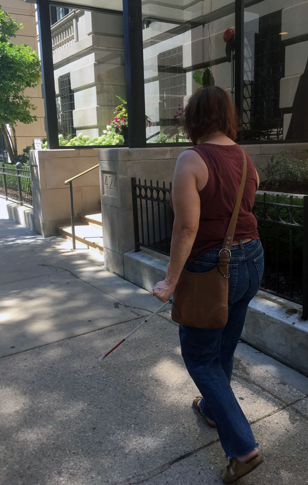 Brooke walks along a city sidewalk near her home using her white cane.